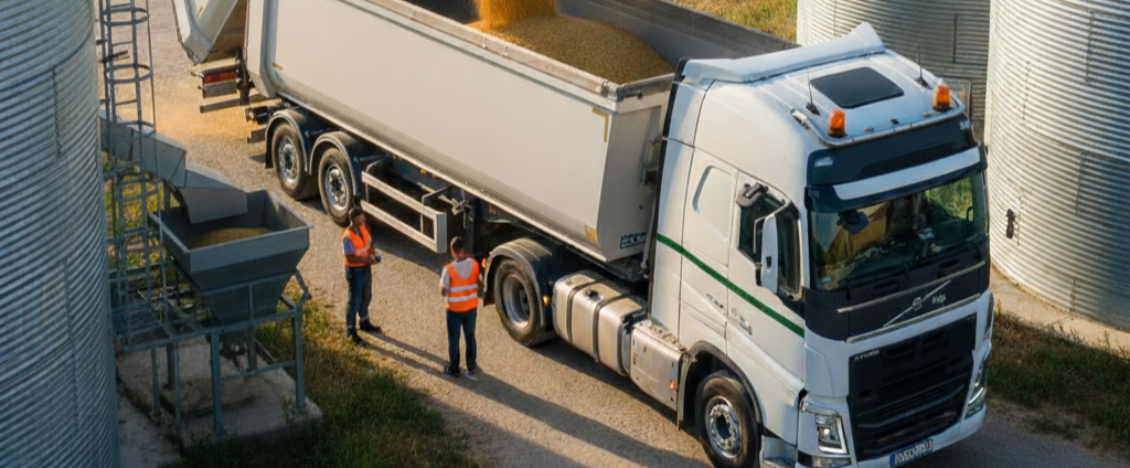 Ciężarówka z naczepą typu wywrotka lub chłodnia na tle pola/silosów, symbolizująca transport zboża lub owoców i warzyw